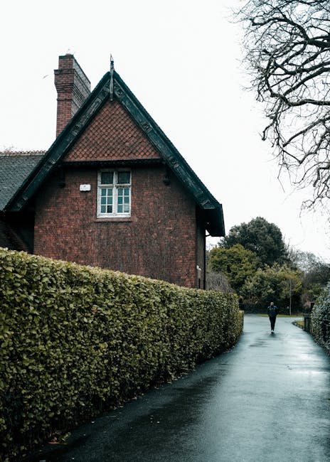 A traditional brick house with a sloped, weathered slate roof and multiple brick chimneys, situated behind a well-maintained garden bed filled with vibrant pink and purple tulips and dark crimson prim