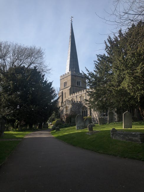 A church with a tall, pointed spire rising prominently against a partly cloudy sky, surrounded by a well-maintained graveyard with weathered headstones. The foreground features a paved pathway flanked