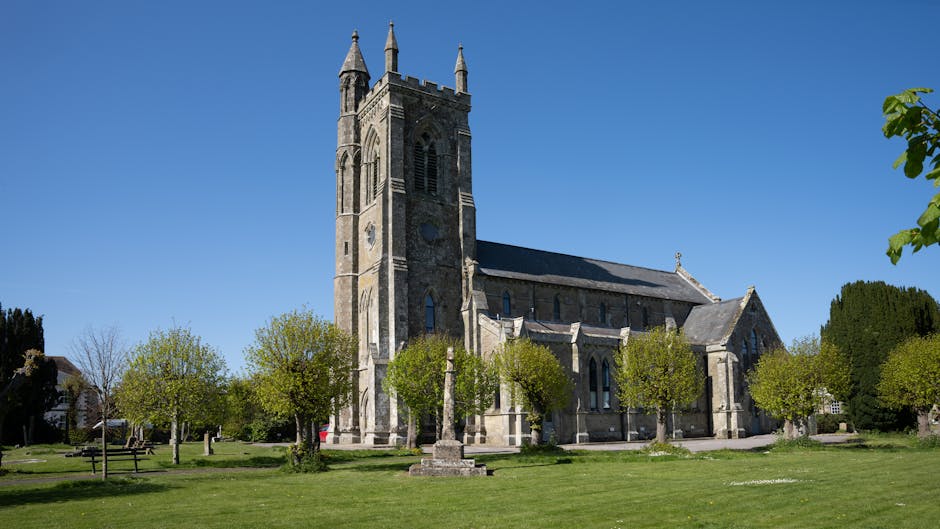A church with a tall, pointed spire rising prominently against a partly cloudy sky, surrounded by a well-maintained graveyard with weathered headstones. The foreground features a paved pathway flanked