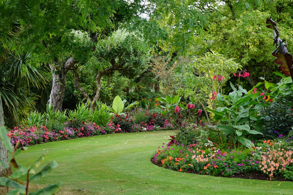 A lush, well-maintained garden scene featuring a curved, neatly mowed lawn bordered by vibrant flower beds filled with pink, purple, orange, and white blooms. Tall, leafy trees with textured bark and 