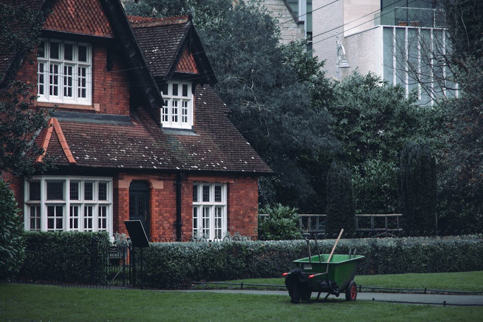 A lush, well-maintained garden scene featuring a curved, neatly mowed lawn bordered by vibrant flower beds filled with pink, purple, orange, and white blooms. Tall, leafy trees with textured bark and 
